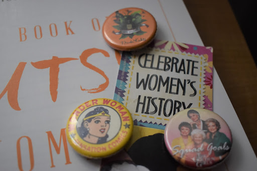 Pins featuring prominent women in pop culture frame a bookmark declaring “Celebrate Women’s History” in honor of Women’s History Month.
