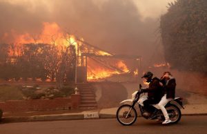 Two people hurry to get past a burning house in the Pacific Palisades neighborhood on Tuesday, January 7th, 2024.  They are trying to record the destruction to send to their family and friends and post on their social media accounts.
